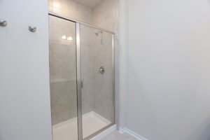Glass shower stall with beige tiles, silver handle, and overhead light fixture in a bathroom.