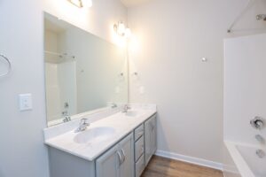 A clean bathroom with a double sink, large mirror, and bathtub on the right.