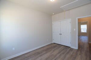 Empty room with wooden floors, white walls, double closet doors, and a small window in the adjacent room.