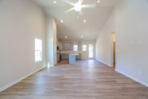 Spacious empty living room with wood flooring, vaulted ceiling, and open kitchen with modern appliances in the background.