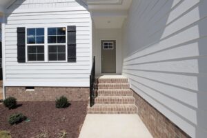 White house entrance with brick steps, black shutters, and a green door. Freshly landscaped with small bushes.