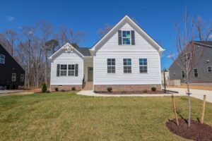 White single-story house with dormer windows, brick foundation, front lawn, and clear blue sky.