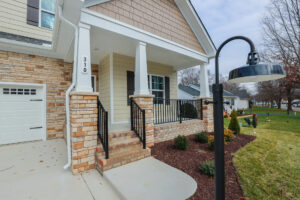 Suburban house entrance with steps, stone columns, and a black lamp post, surrounded by a tidy lawn and shrubs.