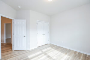 Empty room with white walls, wooden floor, open door, and closed closet doors. Natural light streaming through a window.