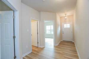 Bright hallway with wooden floors, white walls, and doors leading to rooms. A light fixture hangs from the ceiling.