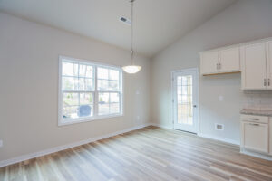 Empty room with wood floors, large window, pendant light, and door leading outside. Kitchen cabinets on the right.