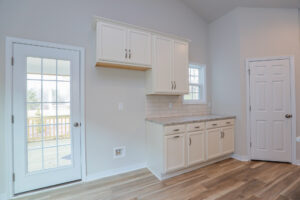 Bright kitchen with white cabinets, granite countertops, wooden floor, and a door to a deck. Large windows provide light.