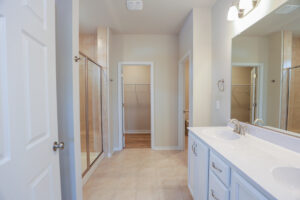 A bright bathroom with a double sink, shower, open closet, and beige tile flooring.