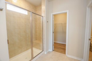 Beige tiled bathroom with a glass shower door, leading to a walk-in closet with wooden flooring and shelves.