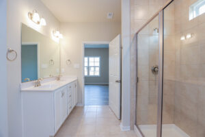 Modern bathroom with double sinks, large mirror, tiled shower, and open door leading to a bright room.