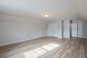 A spacious, empty room with light wood flooring, white walls, a ceiling light, and two white double doors.