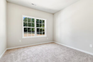 Empty room with beige carpet, light gray walls in Patrick Henry Heights, Lot 57, with a window overlooking trees and grass.