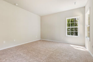 Empty carpeted room with beige walls and sunlight through two windows in Patrick Henry Heights, Lot 57.