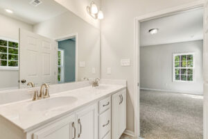 White bathroom with double sinks, large mirror, and view into Lot 57 at Patrick Henry Heights’ bright, carpeted bedroom.