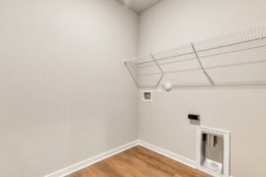 Empty laundry room in Patrick Henry Heights Lot 57 with gray walls, wire shelf, and washer-dryer hookups above wood flooring.