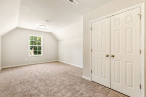 Empty, carpeted room in Patrick Henry Heights’ Lot 57, with sloped ceiling, double closets, and natural light from a window.