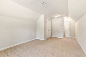 Brightly lit empty room in Lot 57, Patrick Henry Heights with beige walls, carpet, sloped ceiling, and two white doors.