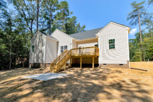 Back view of a white house on Lot 57 in Patrick Henry Heights, featuring a wooden deck and stairs amid trees on a sunny day.