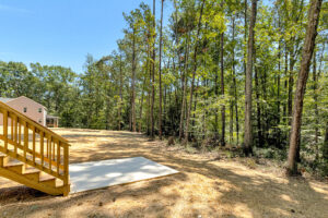 Wooden steps lead to a cleared backyard on Lot 57 in Patrick Henry Heights, bordered by tall trees and blue sky.