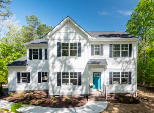 White two-story house with a teal door, large windows, surrounded by trees in Patrick Henry Heights under a clear blue sky.