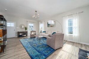 Bright living room in Poplar Village with beige sofas, a blue rug, and modern decor, featuring a large window and chandelier.