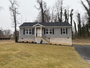 A small, gray house with a porch in Poplar Village, Lot 14, surrounded by bare trees on a cloudy day.
