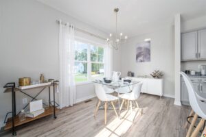 Modern dining area in Poplar Village featuring a glass table, white chairs, chic bar cart, and chandelier by a large window.