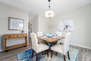 Modern dining room on Lot 22 with a wooden table, white chairs, blue accents, abstract wall art, and a geometric light fixture.