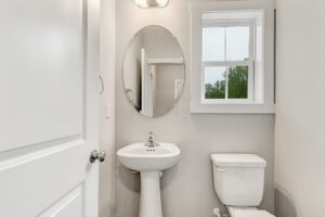 Small bathroom in Lot 22, Poplar Village, with a pedestal sink, round mirror, toilet, and natural light from a window.