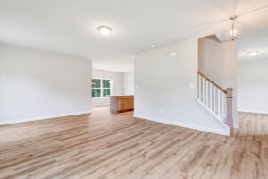 Bright, empty living space in Poplar Village with light wood floors, white walls, and view of kitchen and staircase.