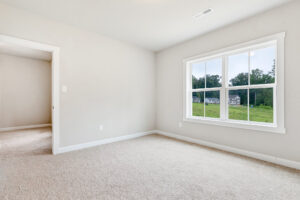 Empty, carpeted room in Poplar Village Lot 22 with large window and doorway overlooking green landscape.