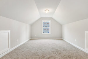Empty, carpeted attic room with sloped ceiling in Poplar Village’s Lot 22, brightened by a single window and neutral walls.