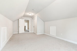 Bright attic room in Poplar Village with sloped ceiling, beige carpet, white walls, and two small side doors.