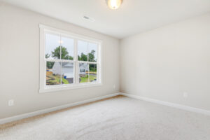 Bright, empty room with beige carpet and large window in Poplar Village’s Lot 22, overlooking a suburban street.