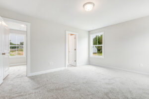Bright, empty carpeted bedroom in Poplar Village Lot 22 with two windows, white walls, and doors to other rooms.