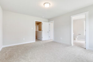Bright, empty bedroom in Poplar Village with beige carpet, white walls, and a doorway leading to a bathroom.