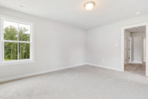 Empty, carpeted room in Poplar Village Lot 22 with white walls, a window showing greenery, and an open hallway doorway.