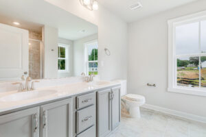 Bright Poplar Village bathroom on Lot 22 with gray cabinets, double sinks, large mirror, and windows with outdoor views.