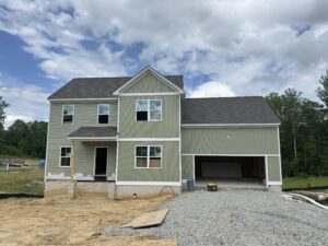 Two-story green house under construction on Lot 22, Poplar Village; garage, dirt, trees, and a cloudy sky above.