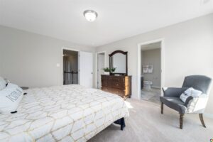 Bright bedroom with a bed, wooden dresser, chair, and open bathroom door in Poplar Villages Lot 22. Neutral tones and natural light.