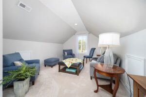 Cozy attic room in Poplar Village with blue chairs, a large window, and a plant. A lamp lights the wooden table and plush carpet.