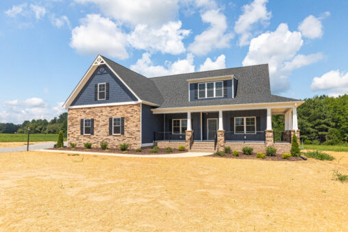 Modern two-story house with blue siding, stone accents, and a covered front porch, set on a sunny, open lot.