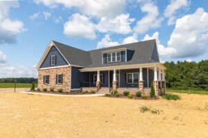 Modern blue house with stone accents and covered porch, surrounded by a dry, grassy landscape under a blue sky.