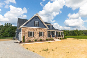 Two-story house with stone and blue siding, large porch, and gravel driveway under a partly cloudy sky.