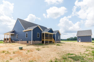 New blue house with covered porch and detached garage on a barren, dirt-filled lot under a partly cloudy sky.