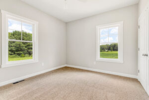 Empty room with beige carpet, two windows, white walls, and a view of greenery outside.