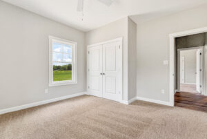Bright, empty bedroom with beige carpet, white closet doors, and a window showing green fields outside.