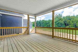 Covered wooden porch with railing, open to a yard and trees under a partly cloudy sky.