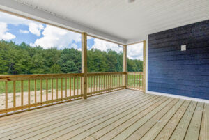 Spacious covered wooden porch with railing, overlooking a grassy yard and forested area under a cloudy sky.