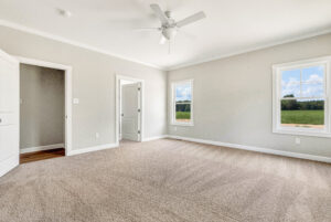 Bright empty bedroom with beige carpet, white walls, two windows, and a ceiling fan.